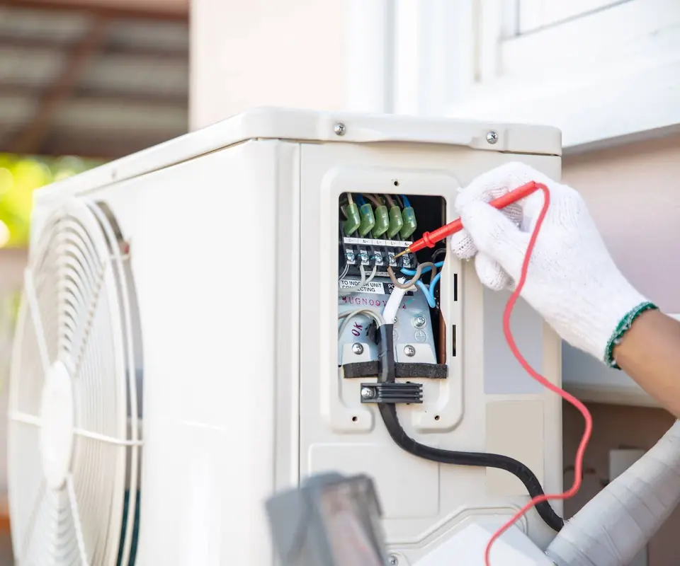 Dłonie w rękawicach sprawdzające miernikiem listwę elektryczną w jednostce zewnętrznej klimatyzacji.