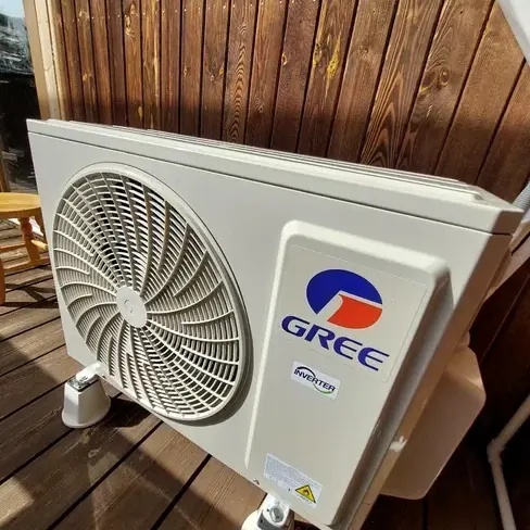 Jednostka zewnętrzna klimatyzacji Gree Inverter zamontowana na drewnianej elewacji domu, ujęcie z dołu na kratkę wentylatora, w tle taras z desek.
