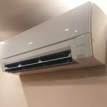 Biała jednostka wewnętrzna klimatyzacji Mitsubishi Electric zamontowana wysoko na ścianie obok narożnika z surowej cegły, w ciepłym świetle punktowym.
