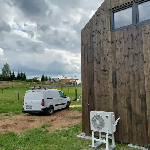 Jednostka zewnętrzna klimatyzacji na stojaku przy ciemnej drewnianej elewacji nowo budowanego domu, obok samochód serwisowy na trawie.