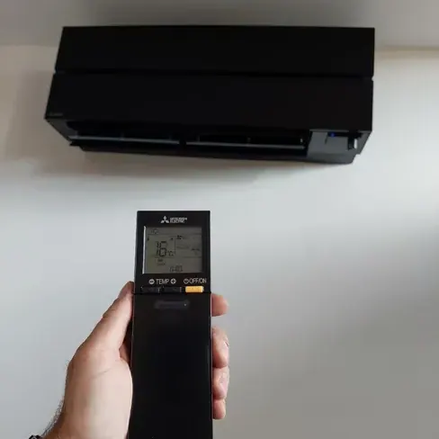 Dłoń trzymająca pilot klimatyzacji Mitsubishi Electric z widocznym wyświetlaczem, w tle czarna jednostka wewnętrzna na ścianie.