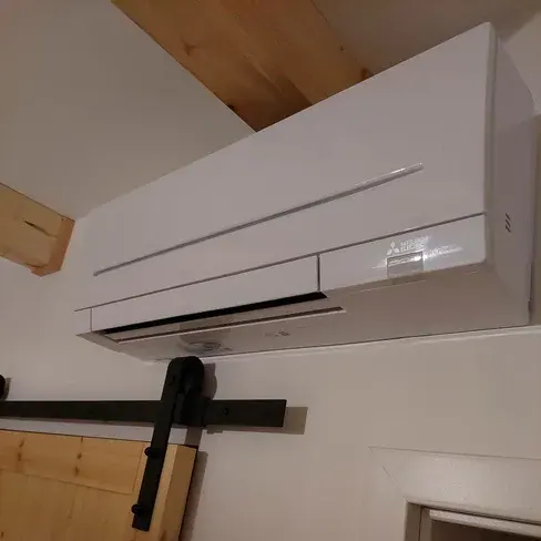 Biała jednostka wewnętrzna klimatyzacji Mitsubishi Electric zamontowana wysoko na ścianie w pomieszczeniu z odsłoniętymi sosnowymi belkami stropu i drewnianymi drzwiami przesuwnymi na czarnym prowadniku.
